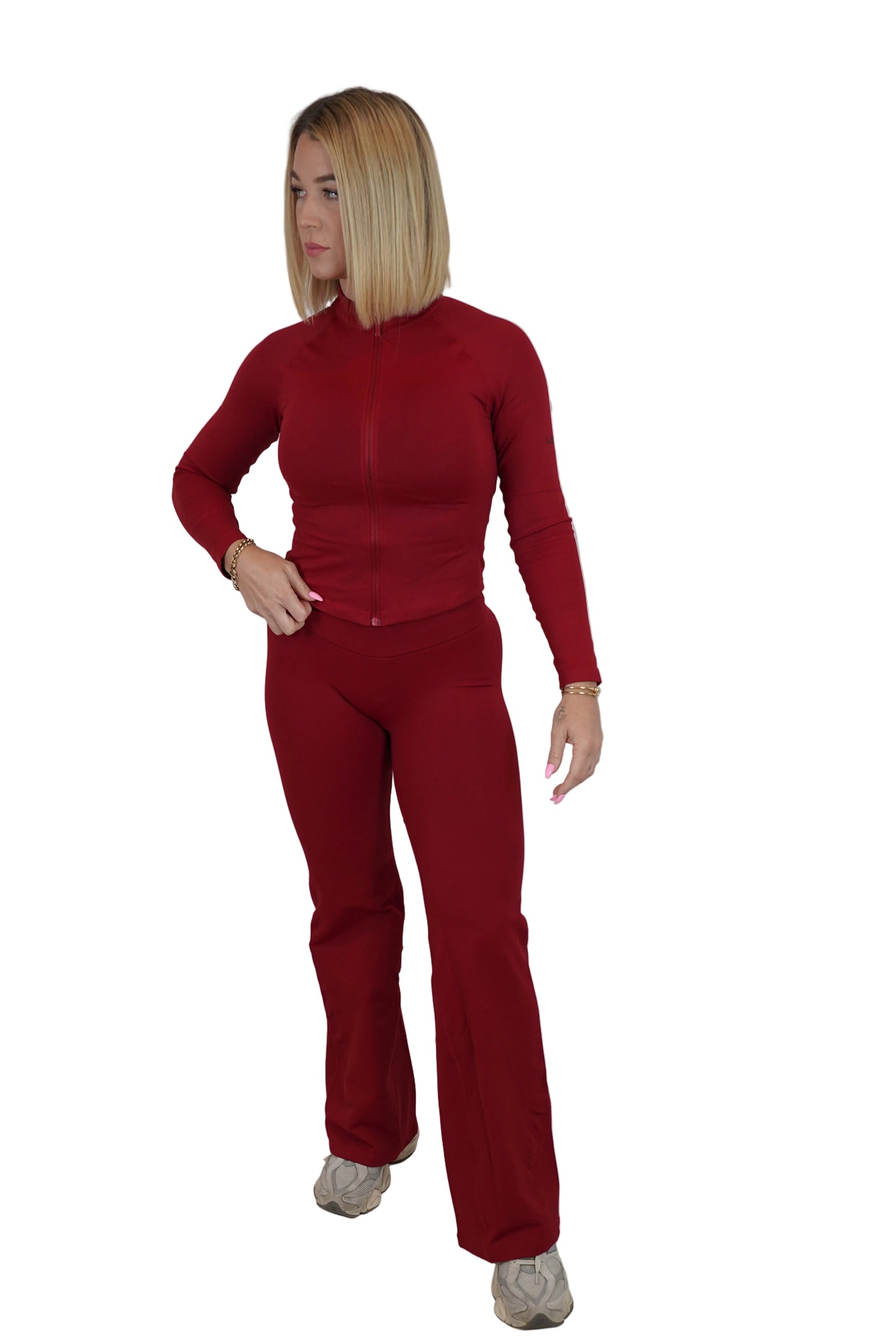 A woman modeling a matching two-piece tracksuit in a deep red color, consisting of a long-sleeve zip-up top and matching pants, standing against a white fabric backdrop.