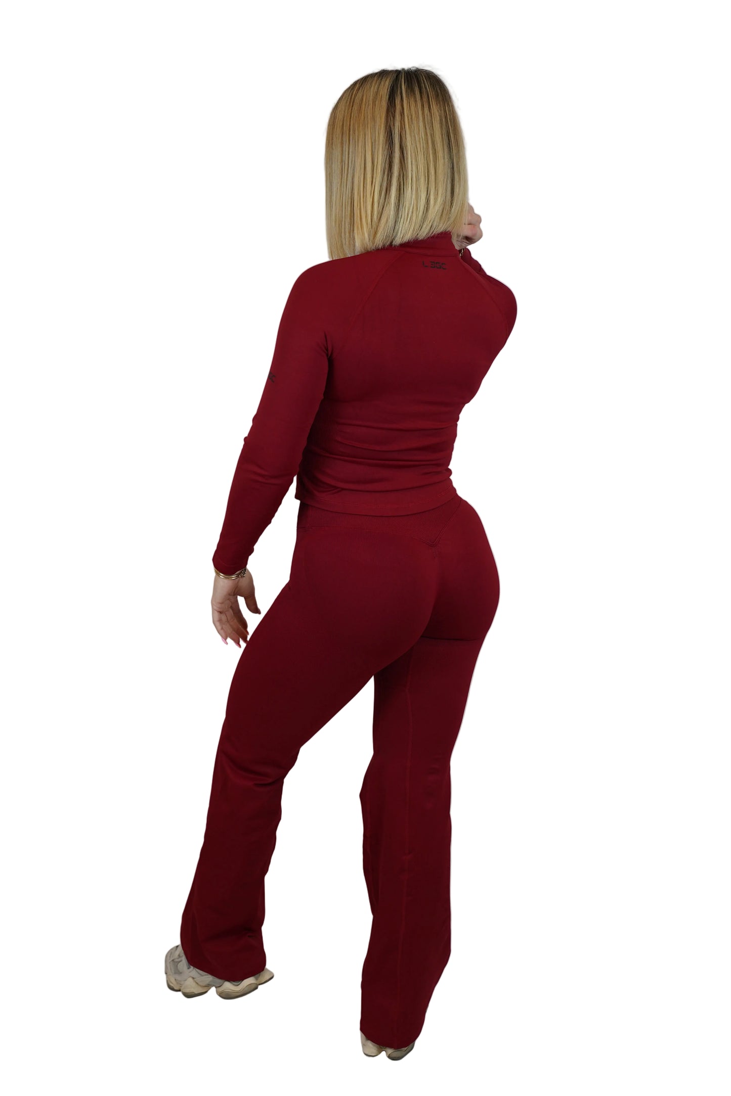 A woman with blonde hair is shown from the back wearing a matching long-sleeve top and flared pants set in a deep burgundy red, standing against a white fabric backdrop.