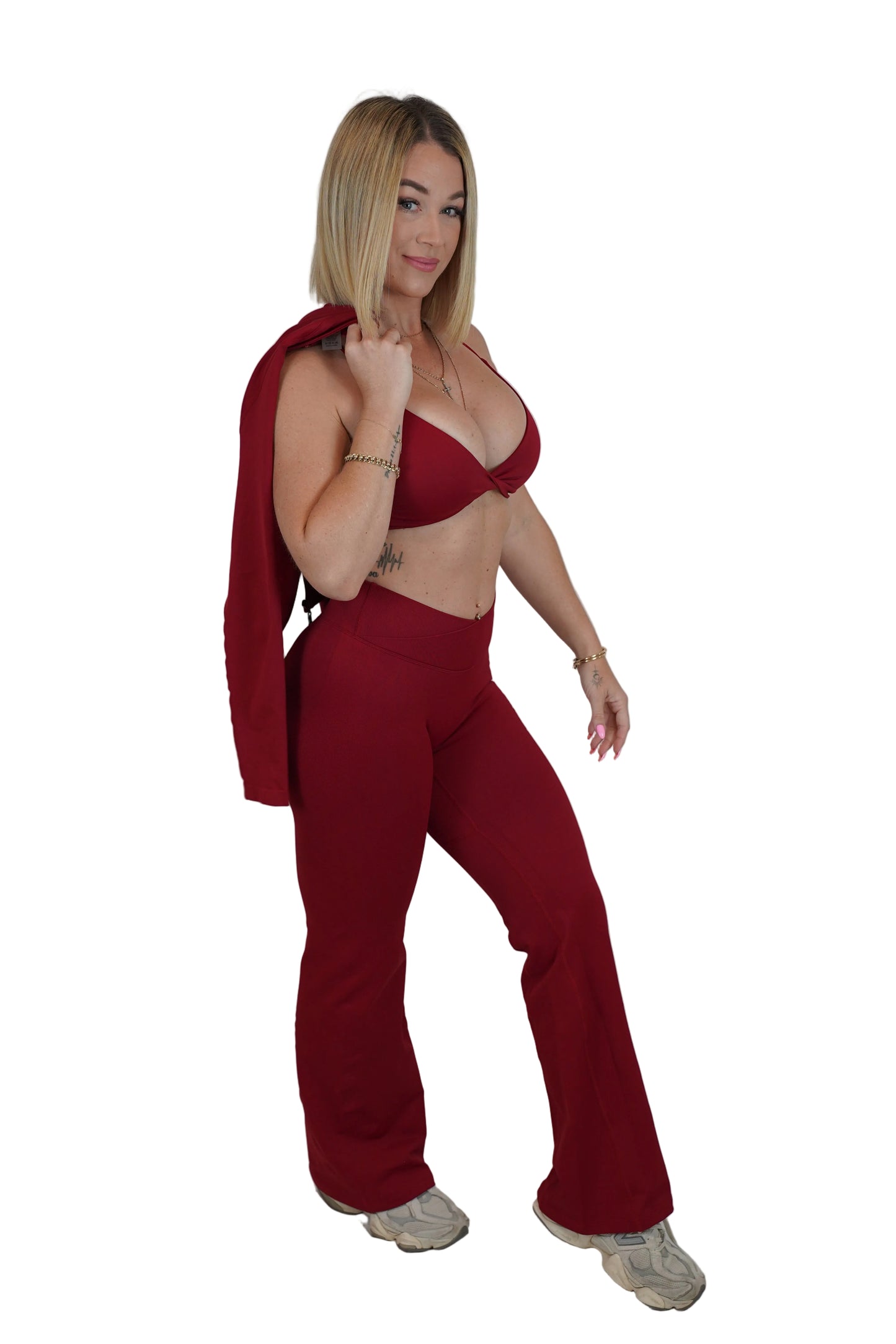 A woman modeling a matching two-piece outfit consisting of a deep red bralette top and high-waisted flared pants, with a red cardigan draped over her shoulder, set against a white studio backdrop.