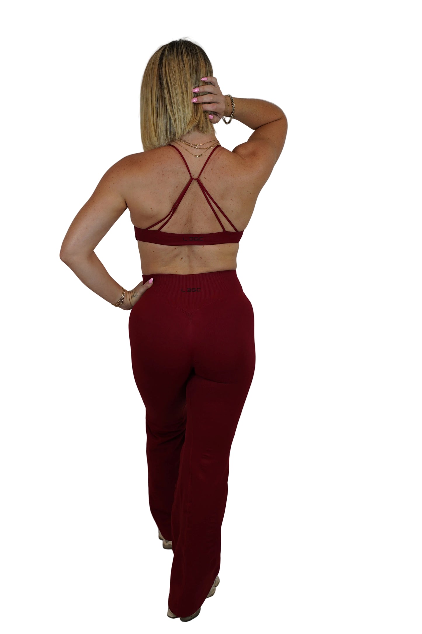 A woman shown from the back wearing a matching burgundy two-piece set consisting of a strappy sports bra and high-waisted pants, standing against a white fabric backdrop.