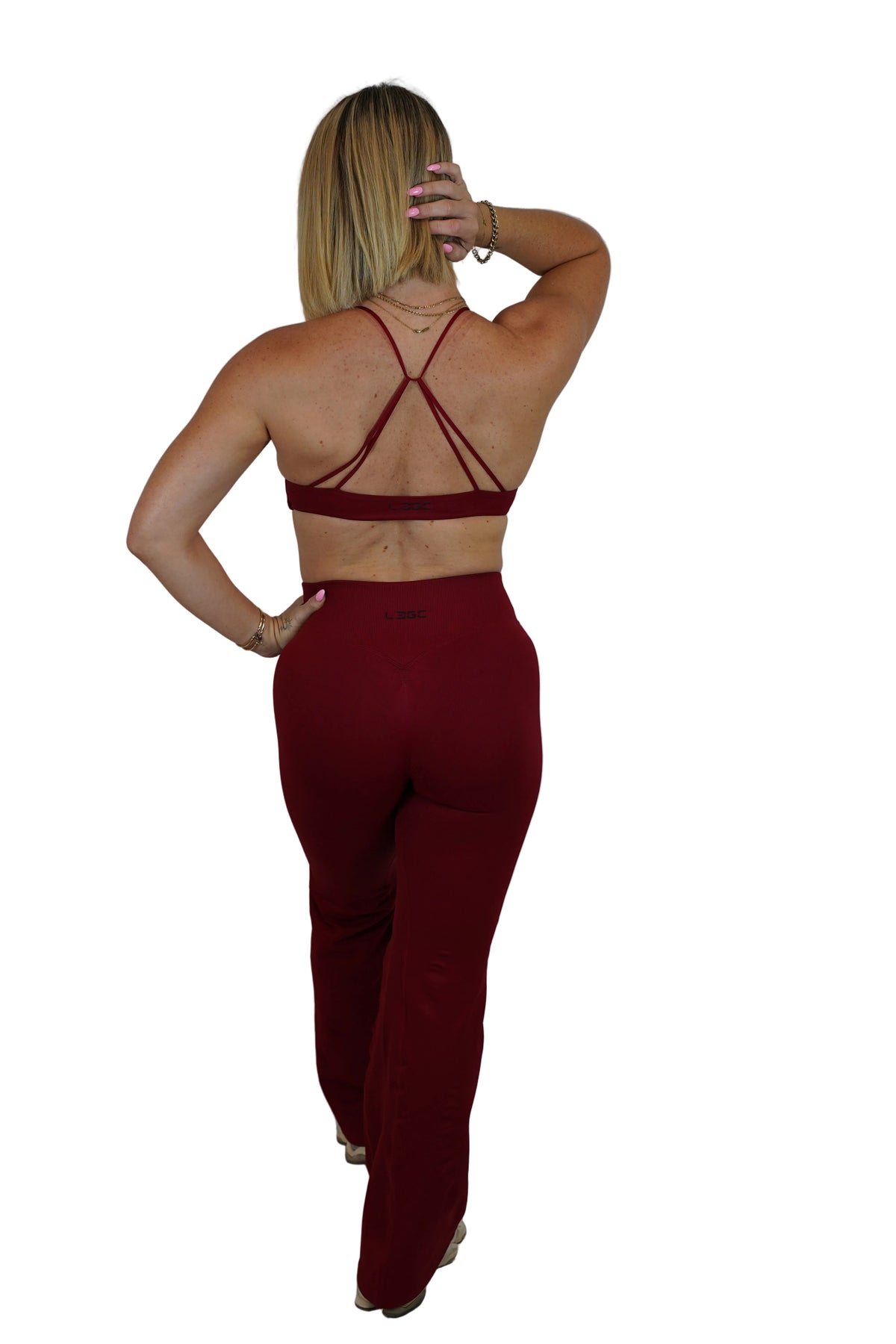 A woman shown from the back wearing a matching burgundy two-piece set consisting of a strappy sports bra and high-waisted pants, standing against a white fabric backdrop.