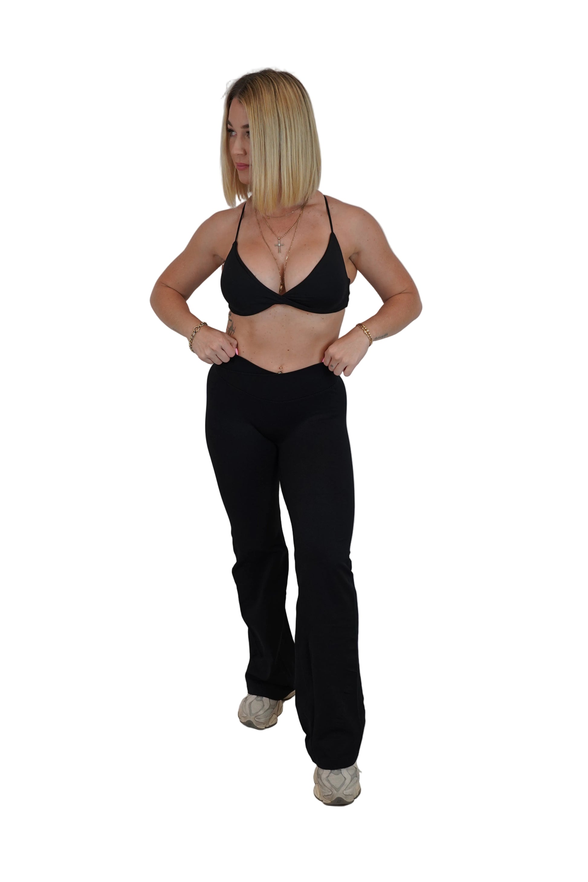 A woman modeling a black two-piece outfit consisting of a triangle bralette and matching high-waisted flared pants, standing against a white fabric backdrop.