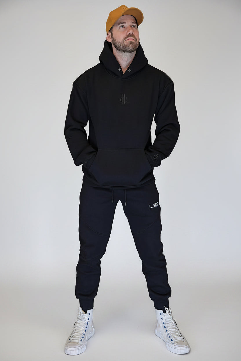 Man wearing a black hoodie, black pants, and white sneakers standing in front of a metallic shutter.