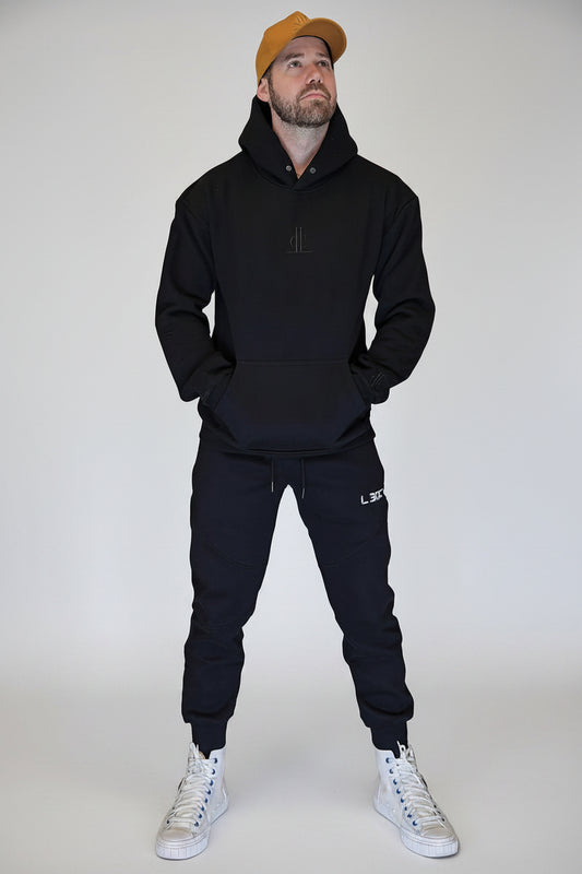 Man wearing a black hoodie, black pants, and white sneakers standing in front of a metallic shutter.