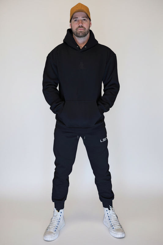 Man wearing a black hoodie and pants with a bronze hat in front of a metallic shutter.