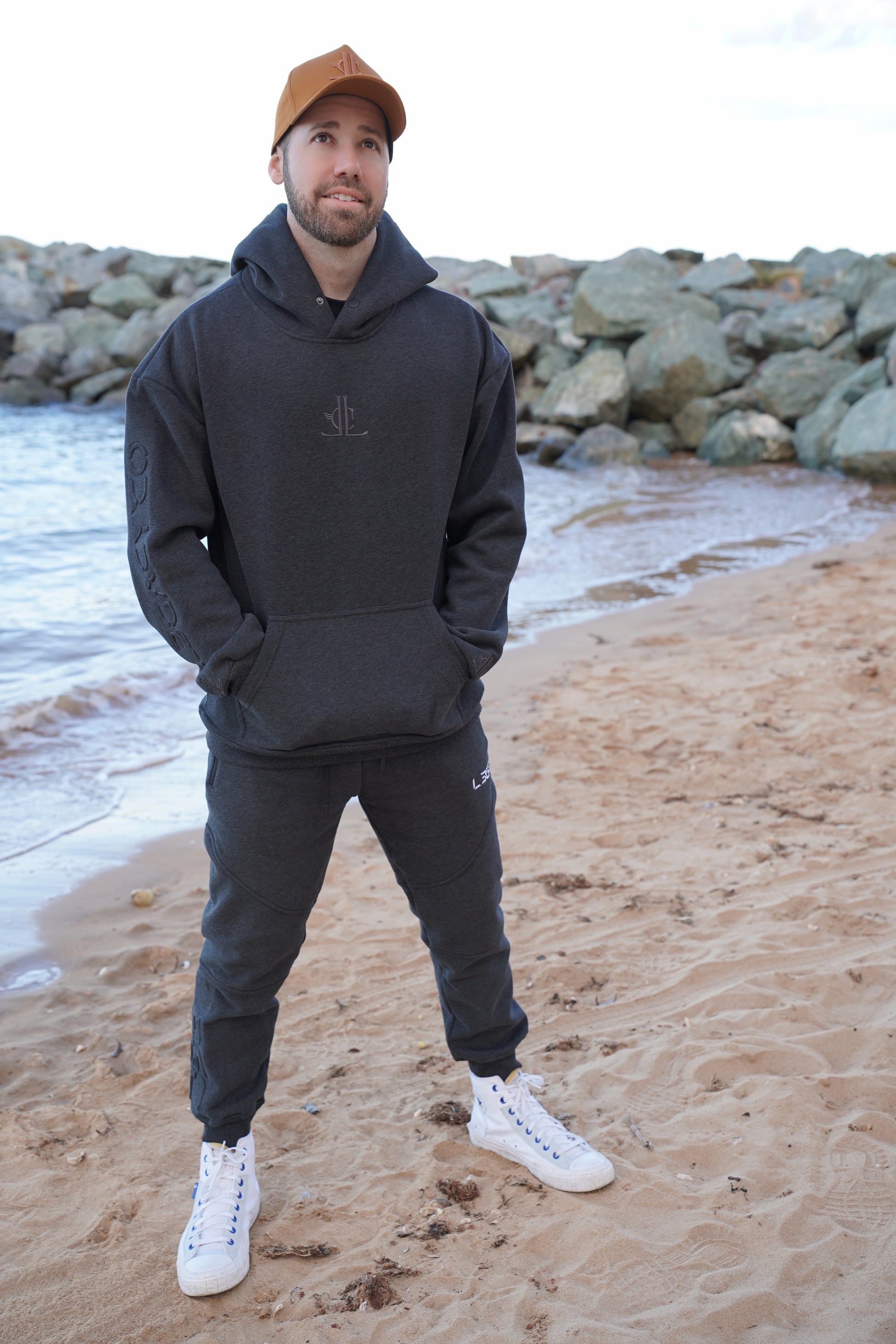 Man wearing a black hoodie and pants with white sneakers on a beach.