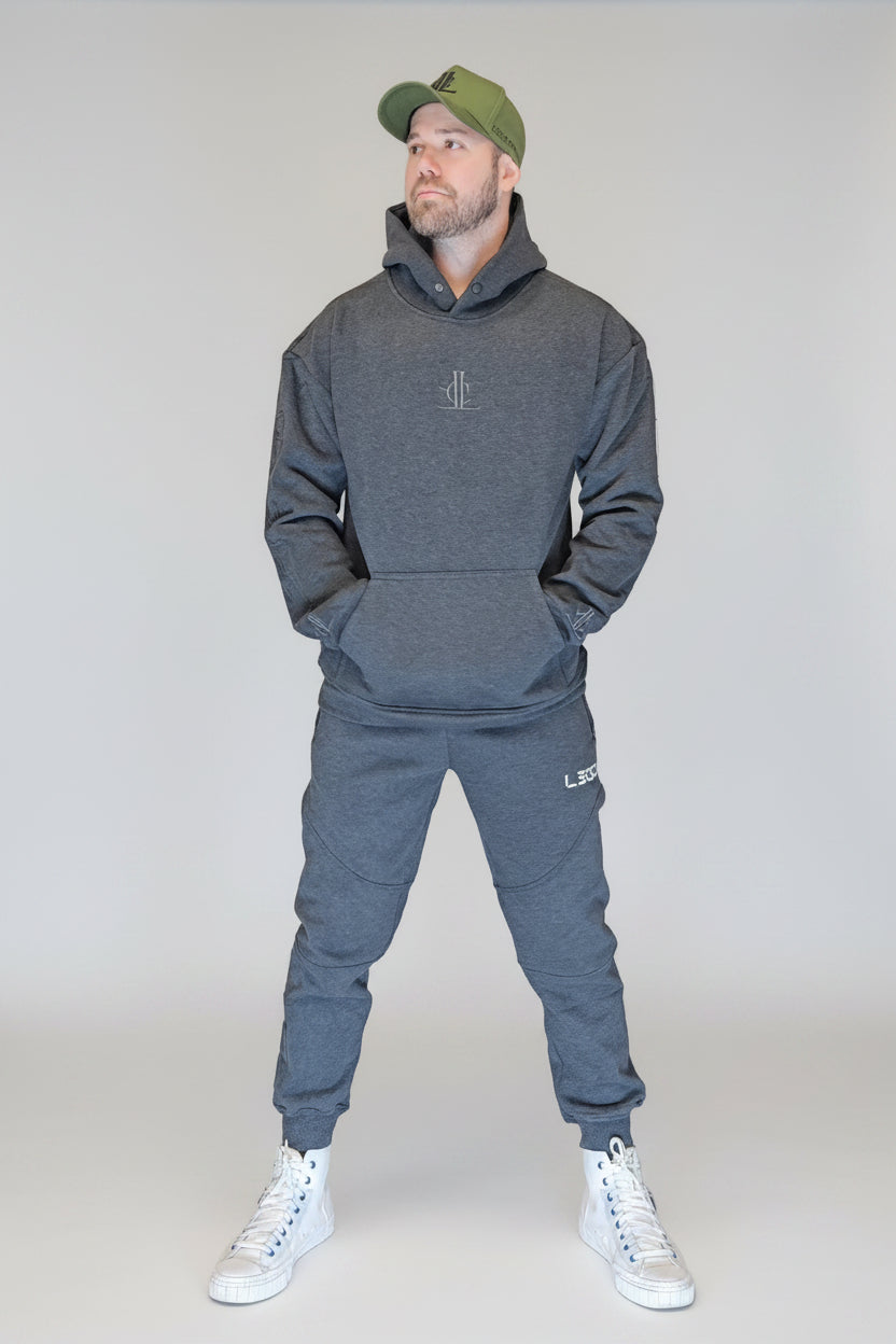 Man wearing a gray hoodie and jogger set with a green cap, standing in front of a metallic shutter.