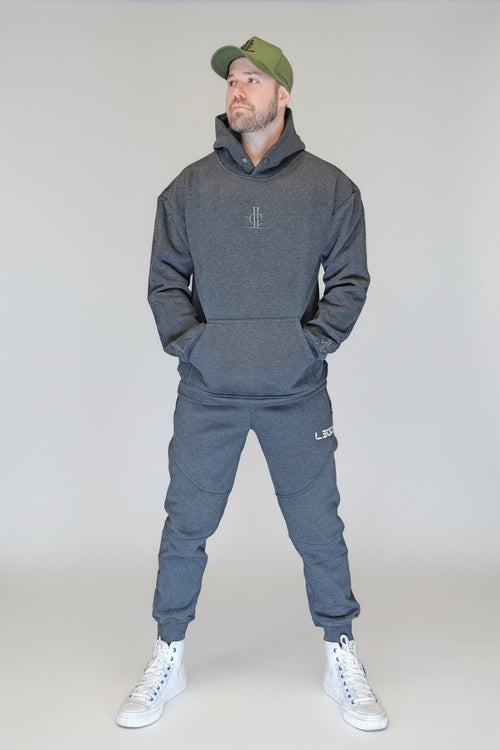 Man wearing a gray hoodie and jogger set with a green cap, standing in front of a metallic shutter.
