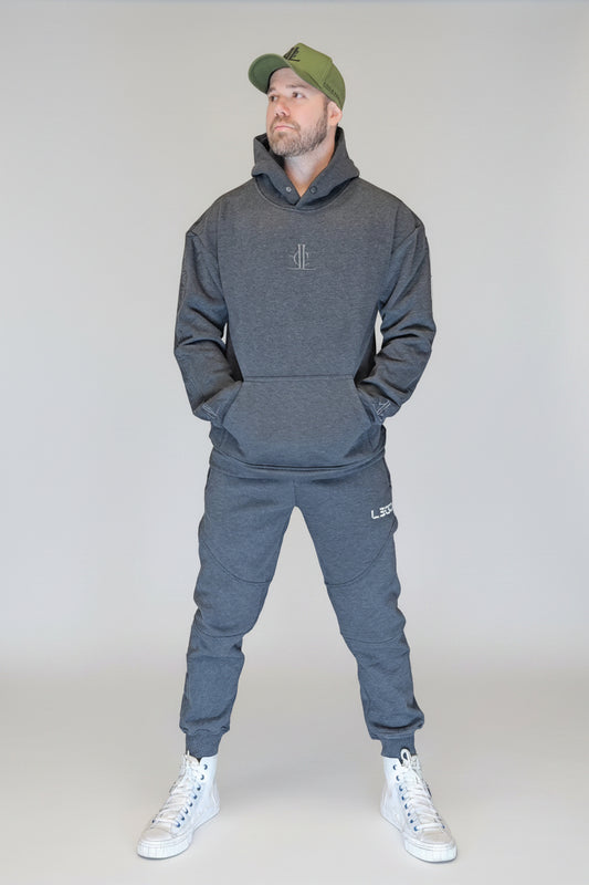 Man wearing a gray hoodie and jogger set with a green cap, standing in front of a metallic shutter.